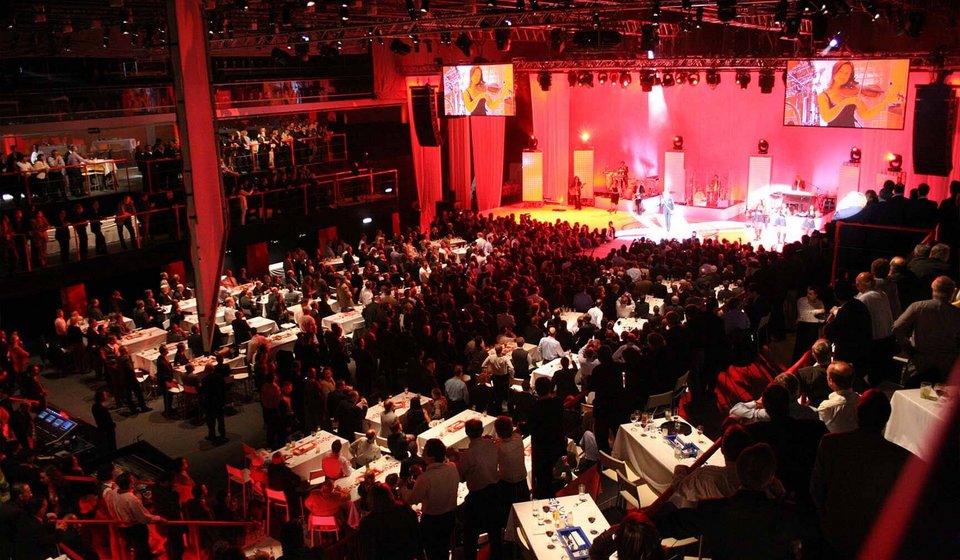 Im Innenraum sind mehrere Dinner-Tische aufgebaut. Die Bühne ist rot beleuchtet. Rechts und links von der Bühne sieht man Übertragungsbildschirme, die eine Geigenspielerin zeigen. Die Saalbeleuchtung ist gedimmt. 